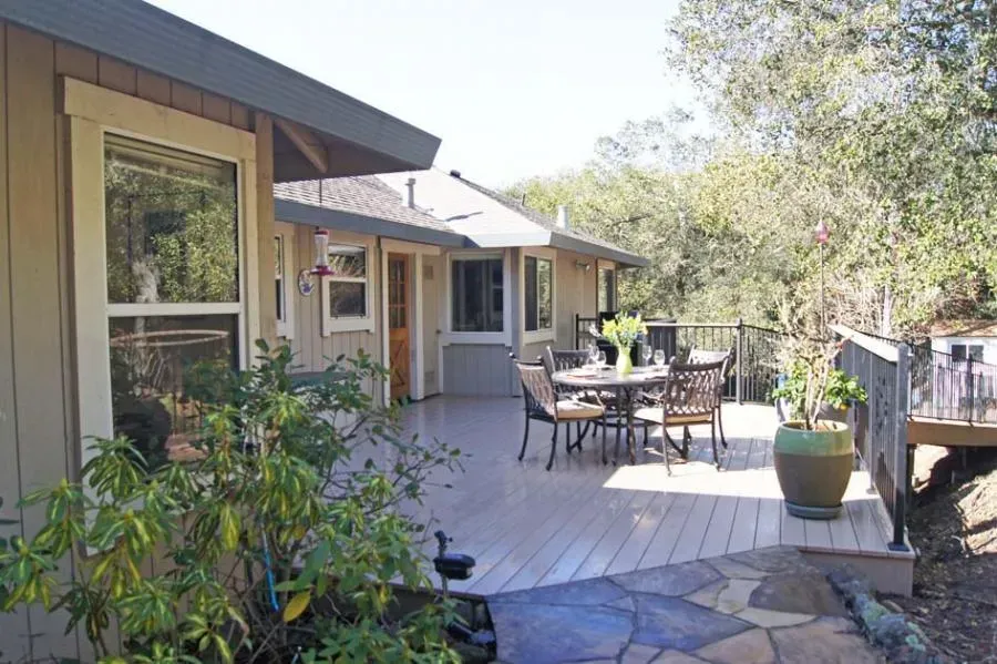 A spacious wooden deck attached to a house, featuring outdoor dining furniture and surrounded by lush greenery.