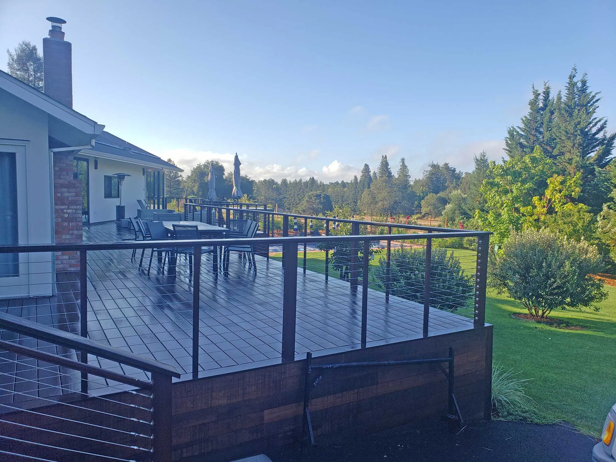 A spacious, modern wooden deck with metal railings and outdoor furniture, set against a backdrop of lush greenery and clear skies in Sebastopol, built by Deckmaster Fine Decks, Inc.