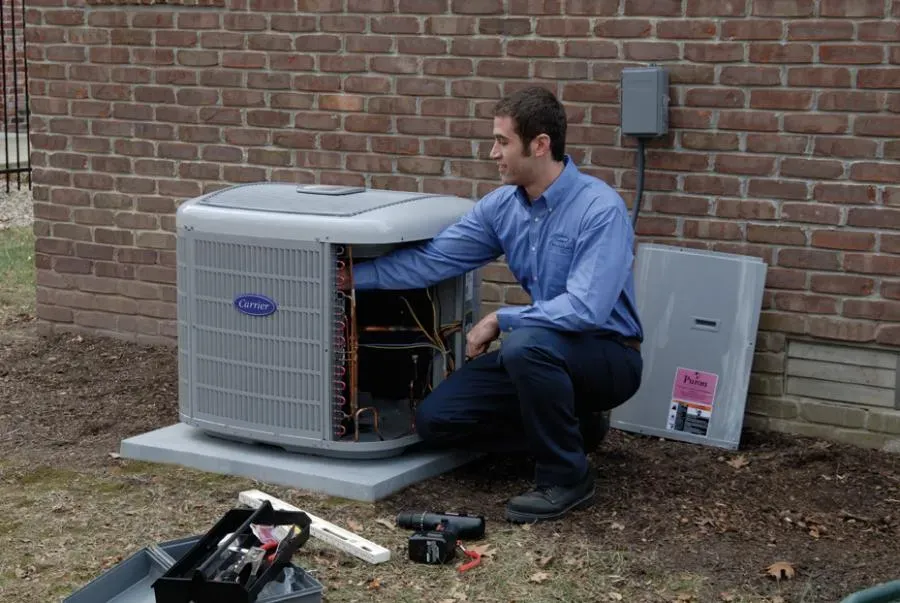 A technician kneels next to an outdoor air conditioning unit, inspecting its components, with a toolbox beside him against a brick wall.