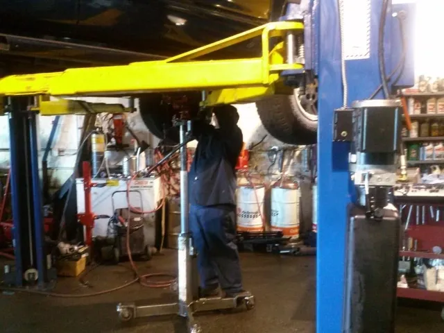 A technician working on a vehicle suspended on a lift at a repair shop, surrounded by automotive equipment and tools.