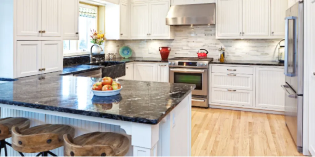 A modern kitchen featuring white cabinets, a large black marble island with wooden stools, stainless steel appliances, and a decorative bowl of fruit on the counter.