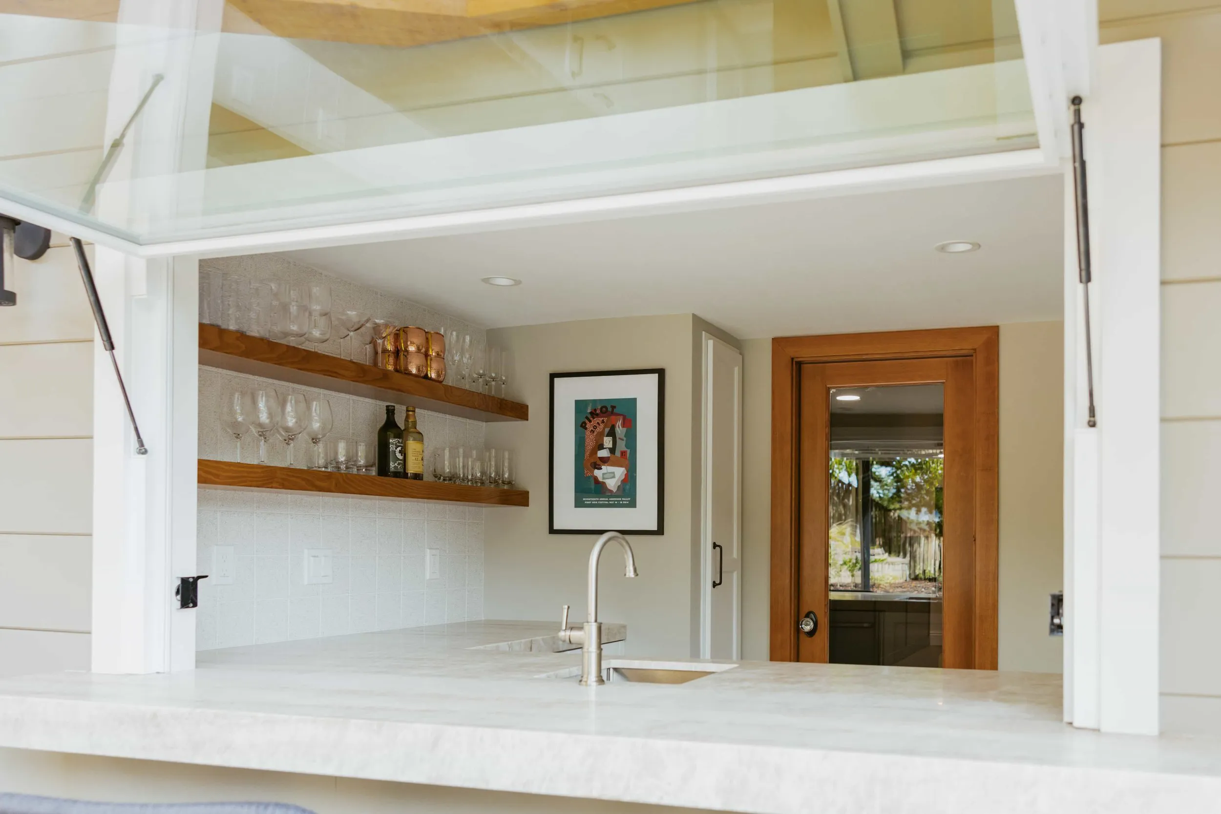 A modern kitchen with a marble countertop, open shelves with glasses and bottles, a stainless steel faucet, and a wooden-framed glass door, designed by Devengenzo & Company in Lafayette.