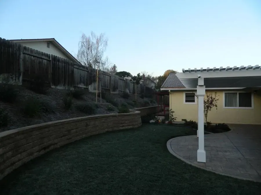 A backyard with a sloping garden, retaining wall, and a patio attached to a yellow house, exemplifying Devengenzo & Company's landscaping design in Lafayette.
