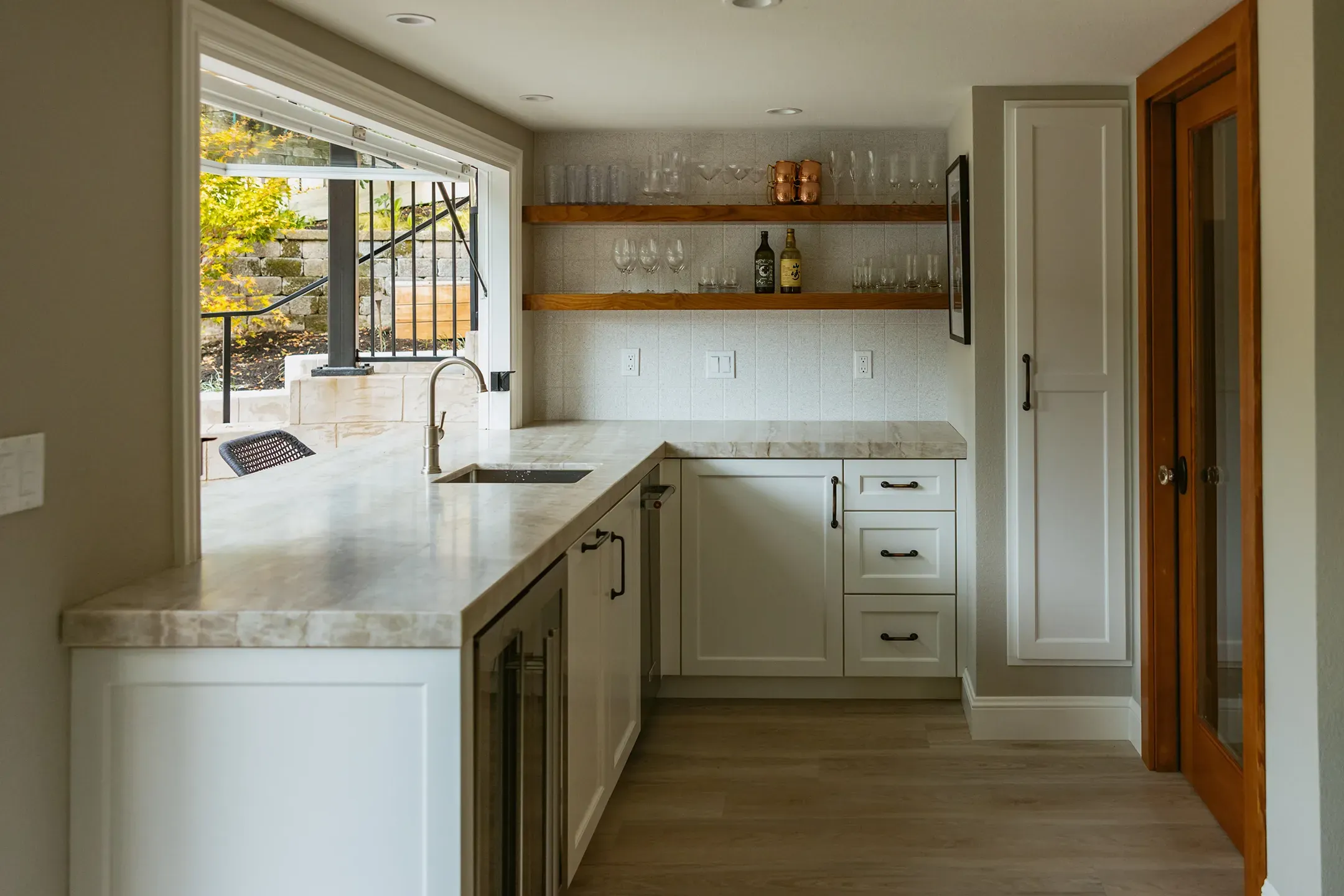 A modern kitchen in Lafayette designed by Devengenzo & Company, featuring white cabinets, marble countertops, wooden shelves with glasses and bottles, and a large open window with a view outside.