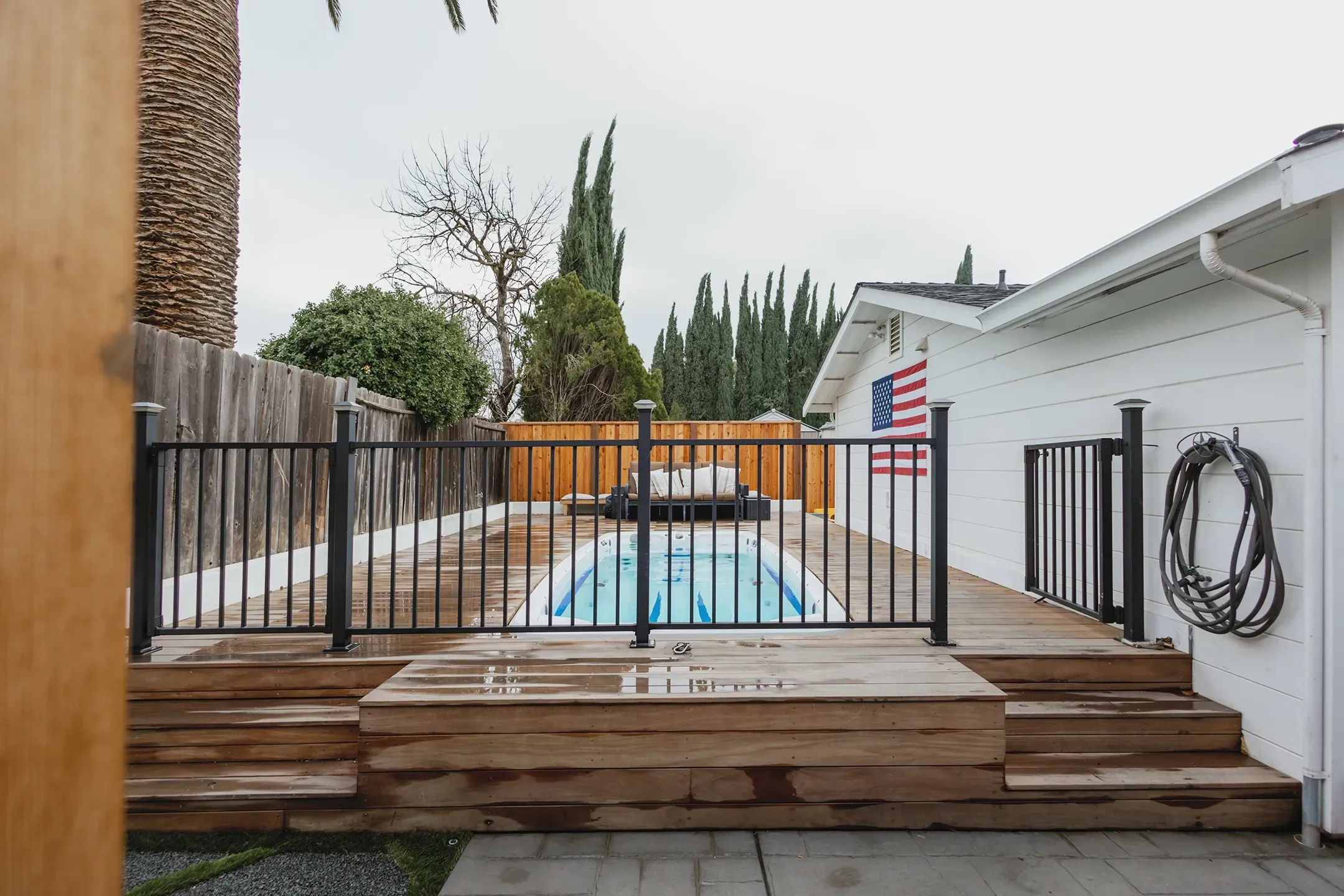 An outdoor pool surrounded by a wooden deck and a black metal fence, with a house displaying an American flag, designed by Devengenzo & Company in Lafayette.