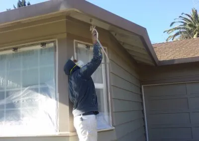 "A person painting the exterior fascia of a house in Santa Rosa, wearing protective clothing, with windows covered for safety as part of DiazandSonsConstruction's service."