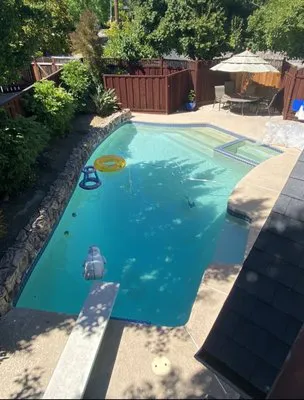 A backyard swimming pool surrounded by a stone border and wooden fencing, featuring a diving board, inflatable pool items, and a seating area with an umbrella.