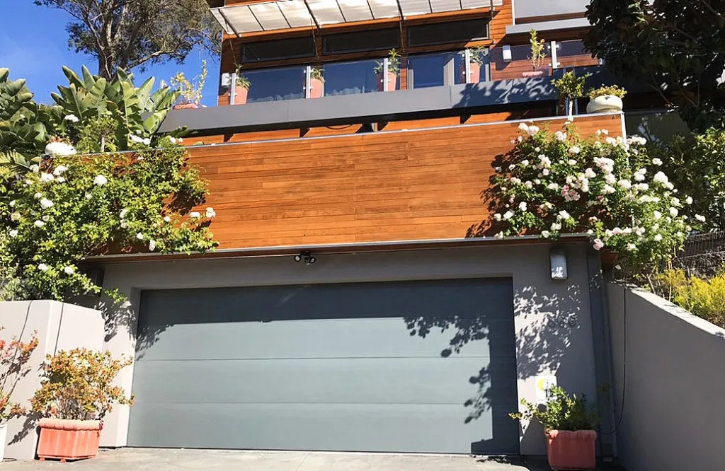 A contemporary house in San Rafael featuring a sleek, gray garage door installed by Door Pros, Inc., surrounded by vibrant greenery and flowers.