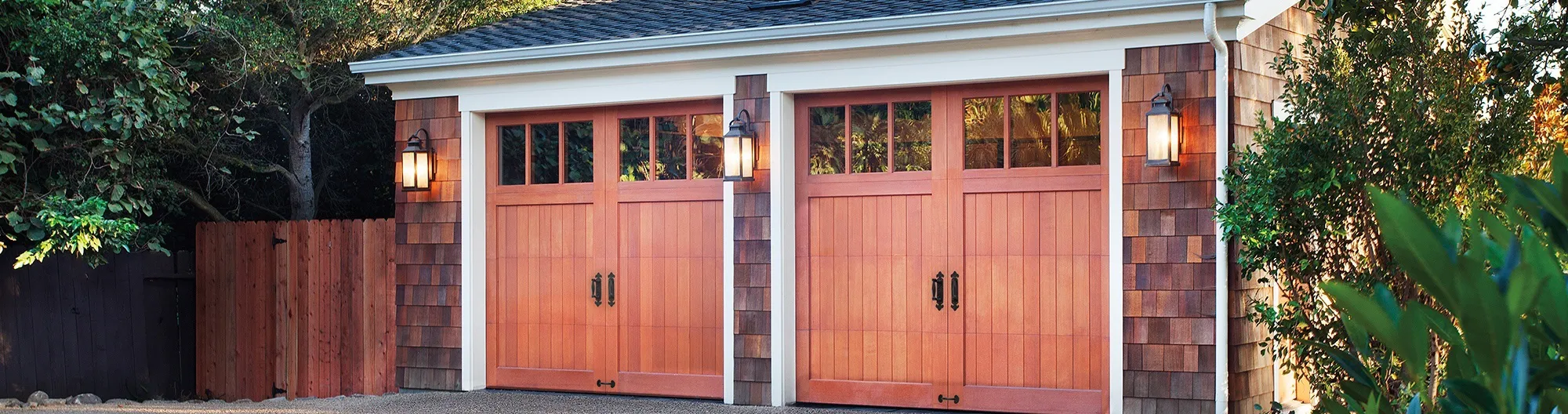 A residential garage with two wooden garage doors, flanked by wall lanterns, surrounded by greenery and a wooden fence.