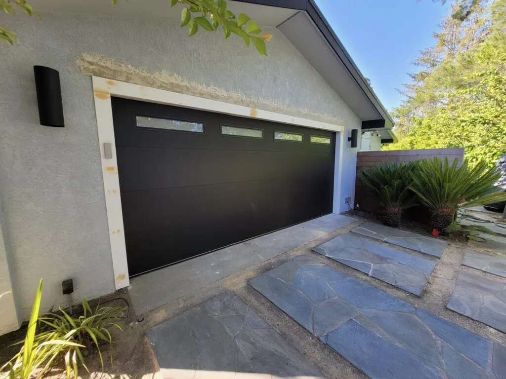 A modern black garage door with rectangular windows, surrounded by a white frame and set against a light-colored house. The driveway features gray stone tiles, and there is greenery on the side.