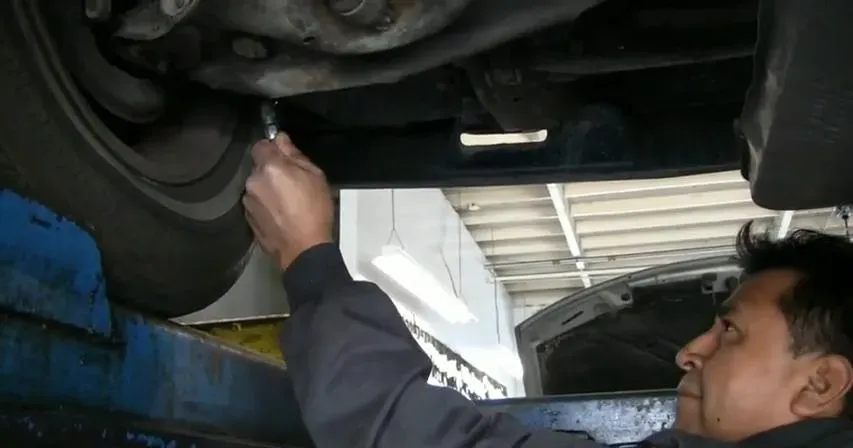 A mechanic working under a car, using a tool to adjust parts, inside a garage with cars and equipment visible.