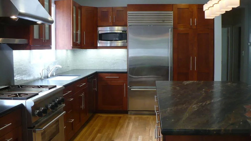 A modern kitchen designed by Dutcher Construction in Sacramento, featuring wooden cabinets, a stainless steel refrigerator, and a marble countertop island.