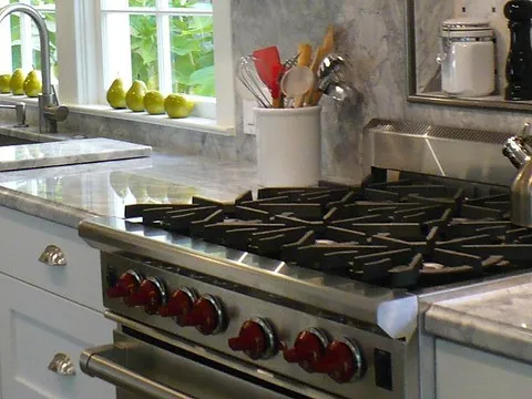 A modern kitchen featuring a stainless steel gas stove with red knobs, marble countertops, a sink, and utensils in a holder by the window.