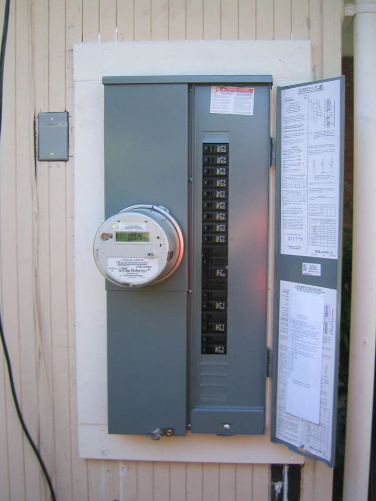 An electrical panel box mounted on a wall with an open door displaying circuit breakers and instruction sheets.
