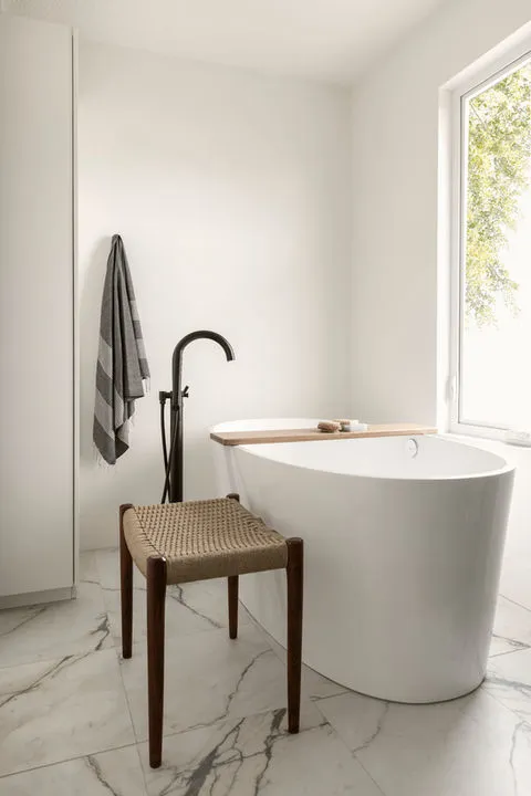 A minimalist bathroom featuring a freestanding white bathtub, a striped towel hanging beside a tall black faucet, and a woven stool on a marble floor, with a window allowing natural light.