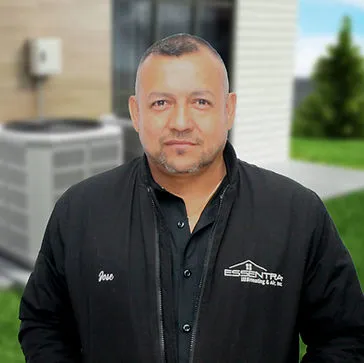 "An individual wearing a black jacket stands in an outdoor setting, with a building and greenery in the background."