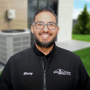 Smiling technician standing outdoors in front of an air conditioning unit, wearing a jacket with company branding visible.