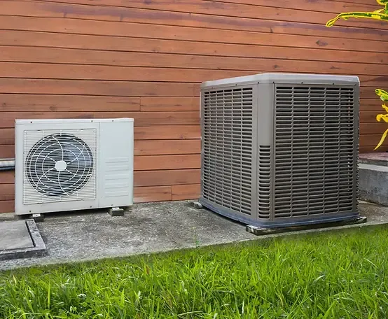 Two outdoor HVAC units positioned on concrete slabs beside a wooden wall, surrounded by green grass.