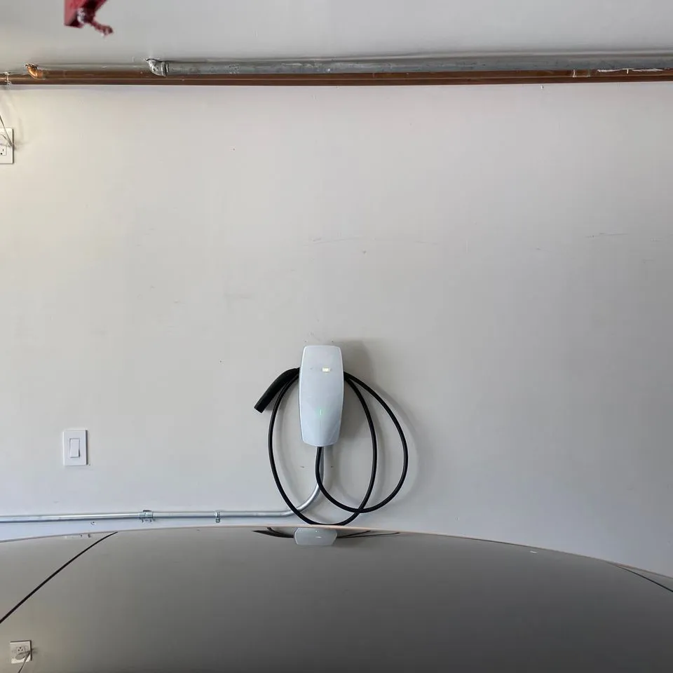 "A white electric vehicle charging station mounted on a wall in a garage, with a car hood visible below, representing Electro-Integrity's home charging setup in San Francisco."
