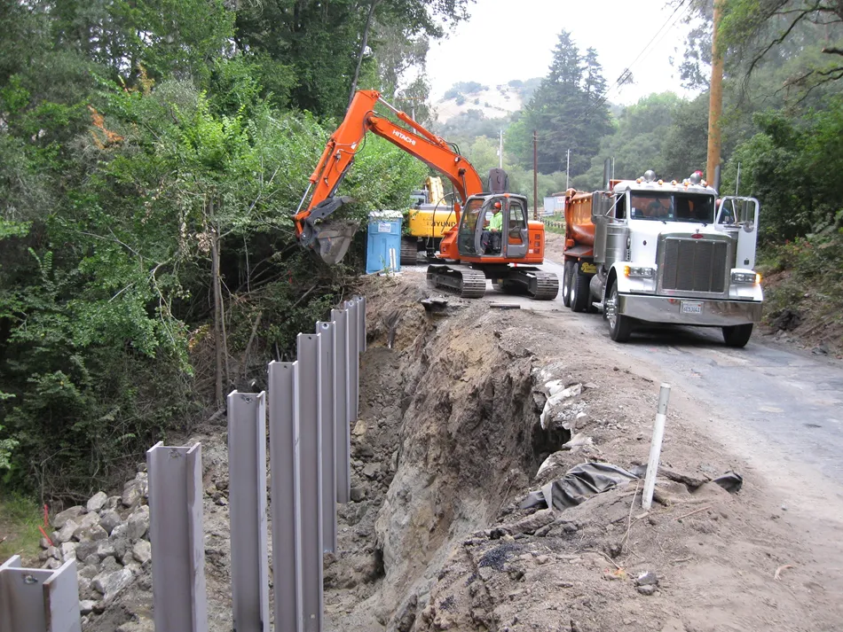 An excavator and a dump truck are positioned on a road alongside a series of installed steel pilings, part of a landslide stabilization project in a wooded area by Engineered Soil Repairs, Inc., in Walnut Creek.