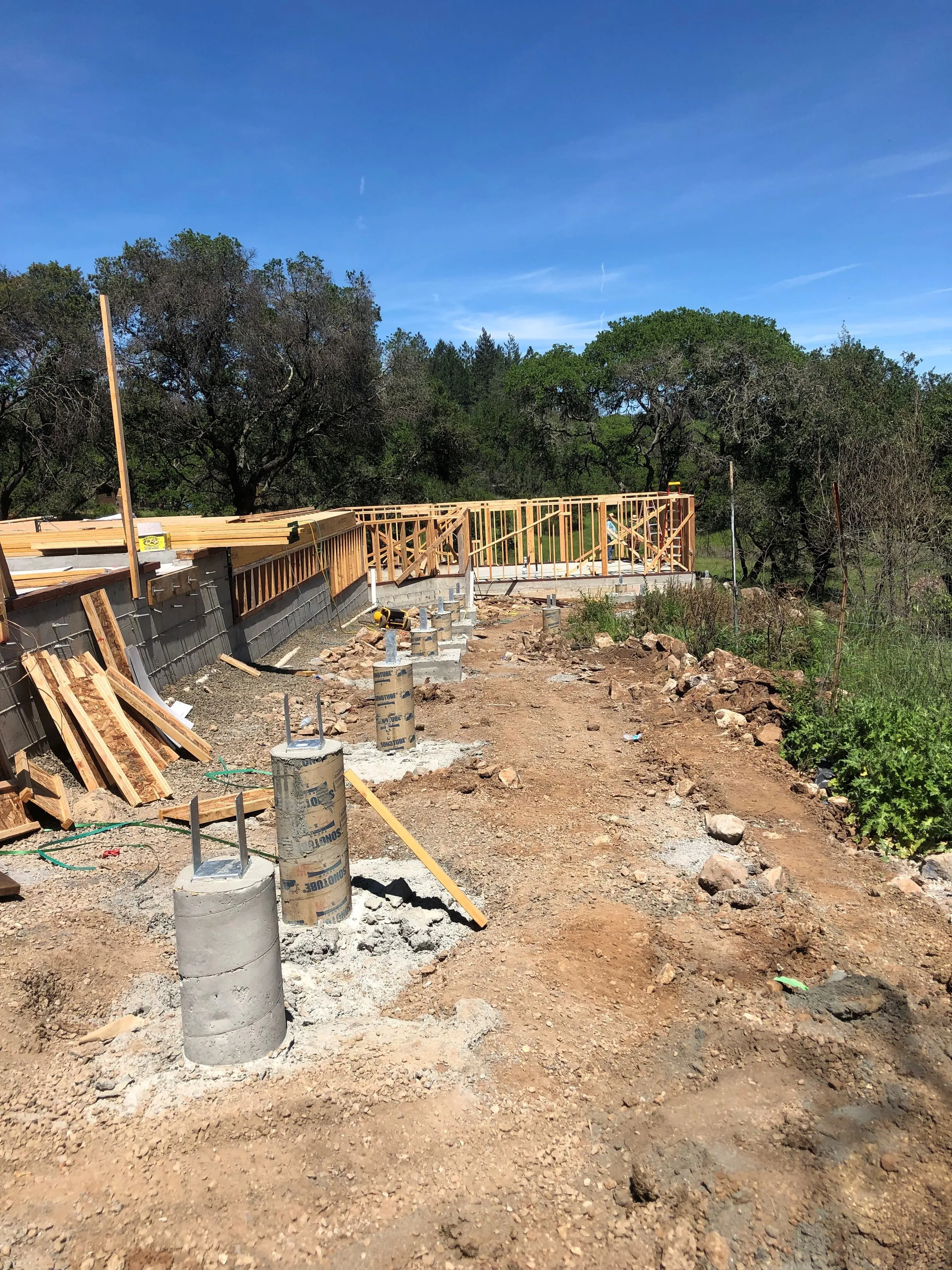 Construction site in Petaluma showing early stages of building by Evenich Construction, Inc., with visible concrete pillars, wooden framework, and surrounding trees.