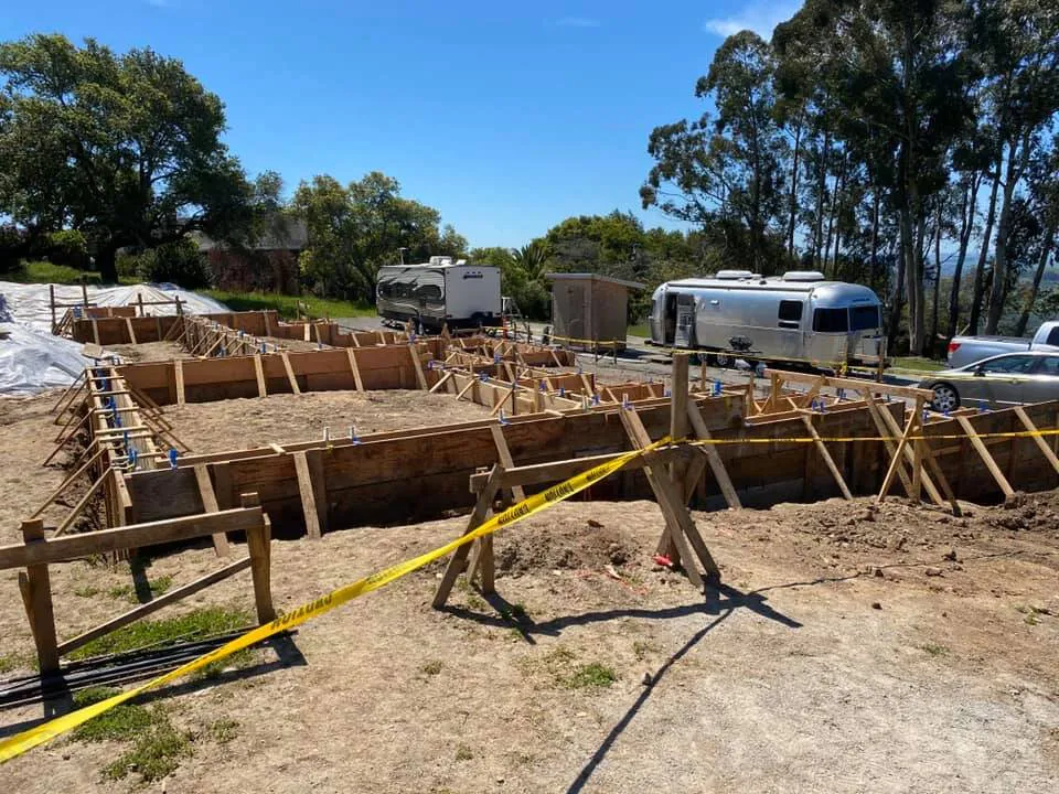 A construction site in Petaluma with wooden framing for a foundation, surrounded by trees and a parked trailer nearby.