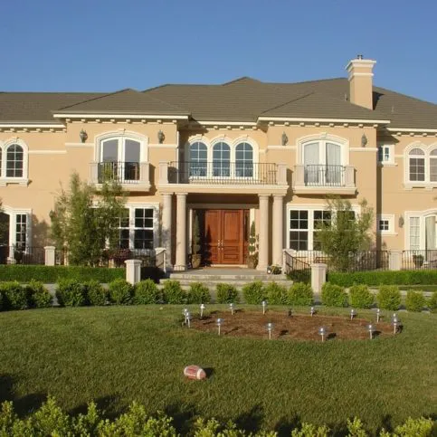 A large, elegant beige mansion with symmetrical windows, a central entrance, manicured lawn, and a small water sprinkler system in front.