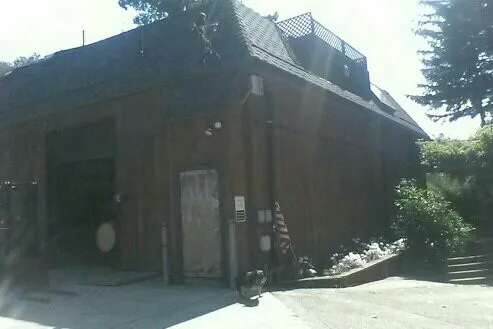 A large wooden building with a dark shingle roof, surrounded by greenery, under bright sunlight.