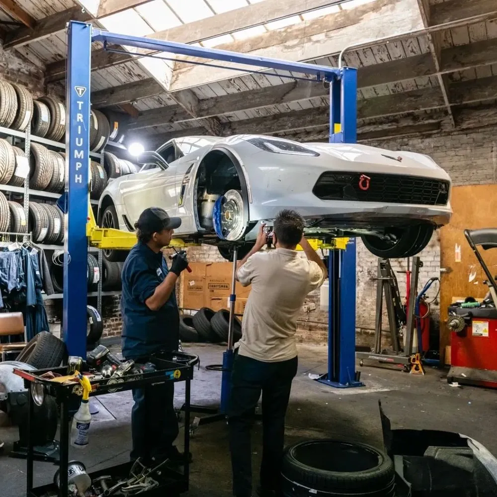 Two mechanics work on a car elevated on a lift in Faxon Garage, San Francisco. The workshop is filled with tires and automotive tools.