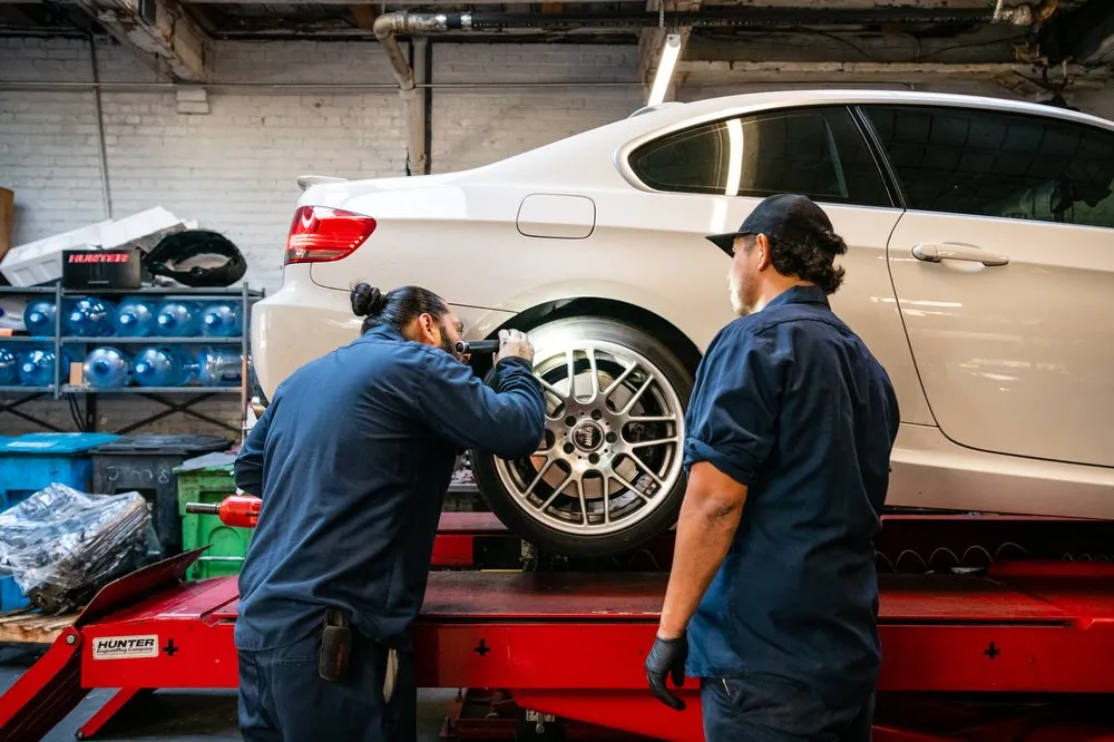 Two mechanics working on a white car elevated on a lift in a garage setting. One mechanic is inspecting a wheel while the other observes.