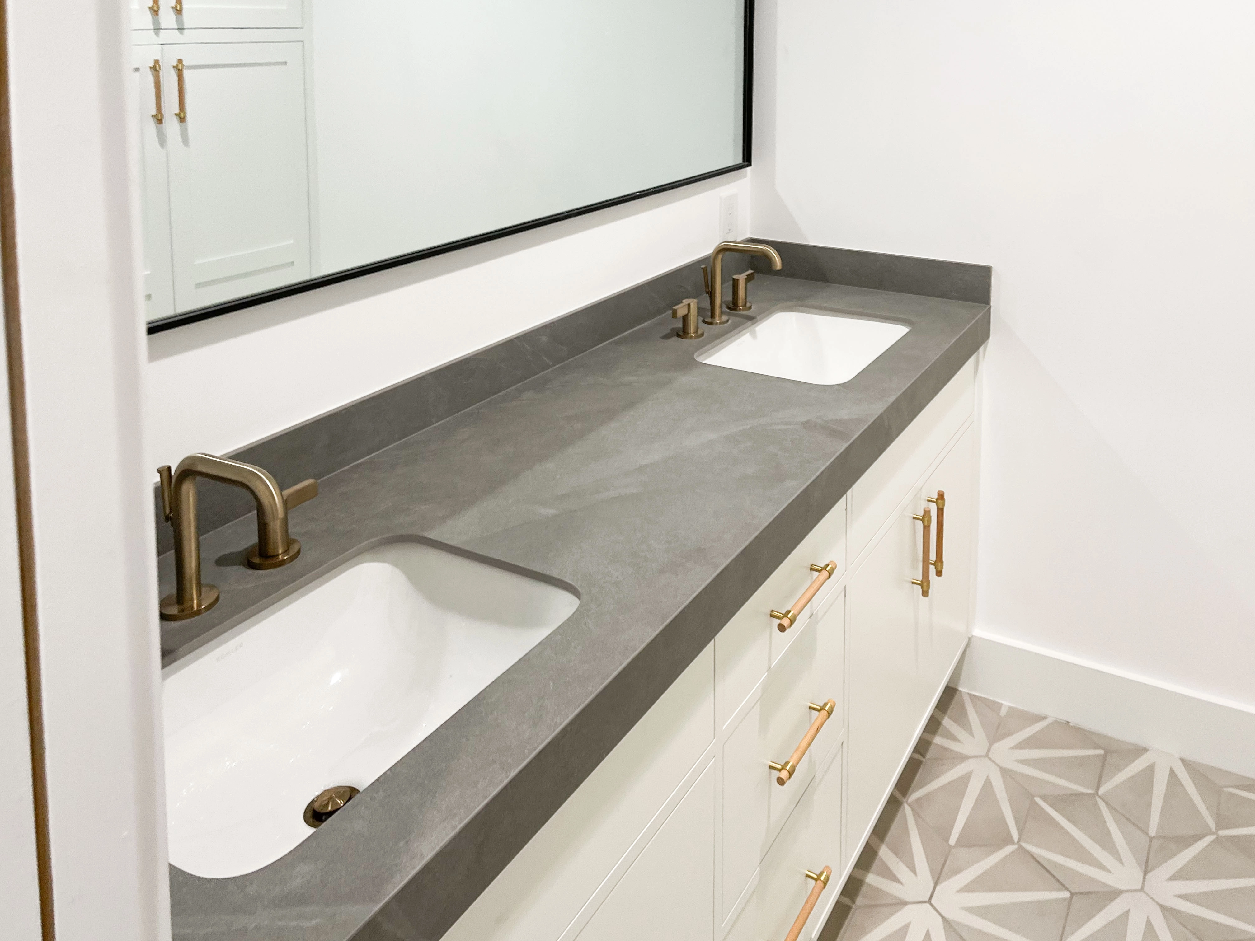 A modern bathroom featuring a double sink vanity with gray countertops, gold faucets, white cabinets, and geometric patterned tile flooring.