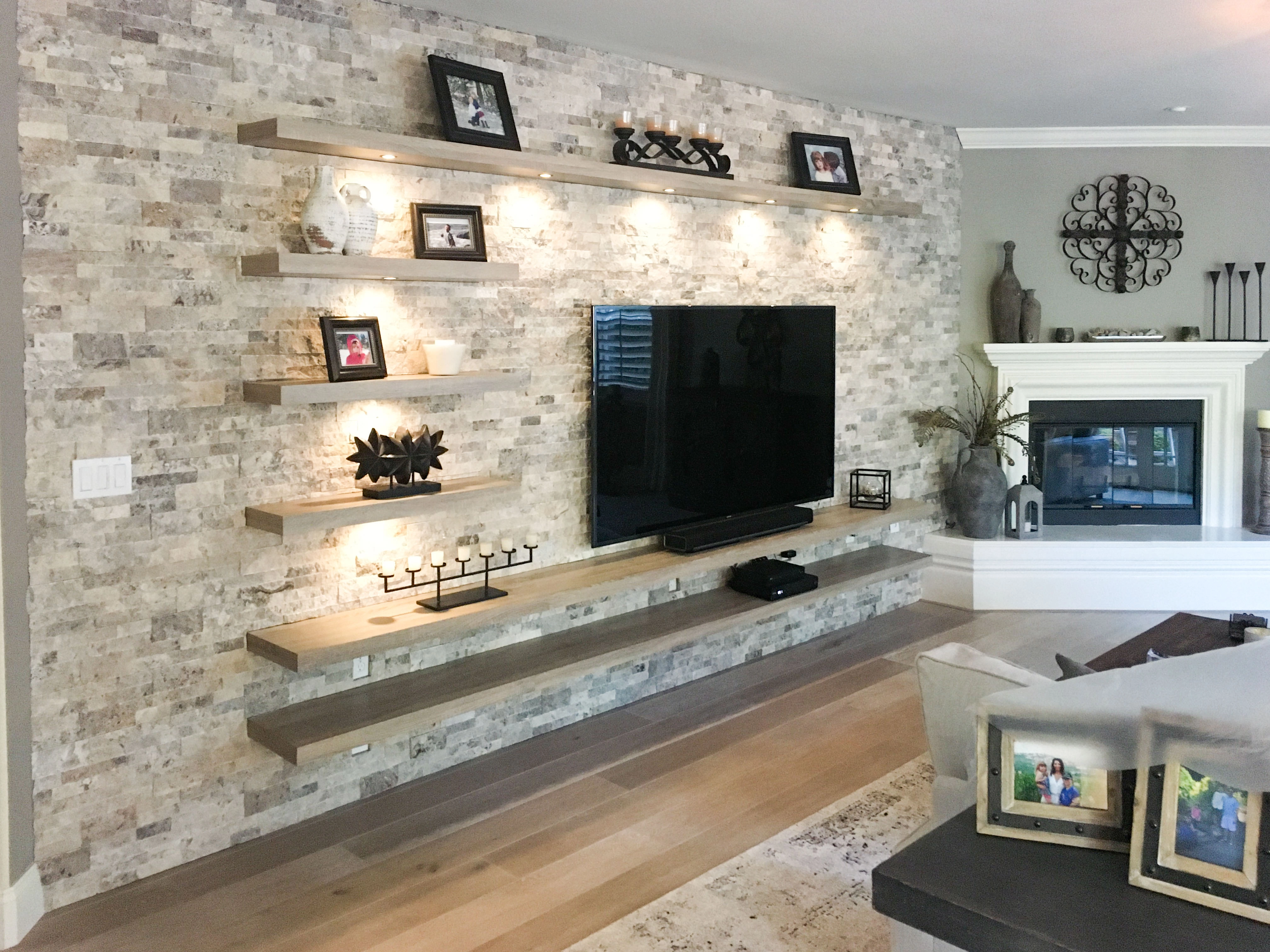 A modern living room featuring a stone accent wall with floating shelves displaying decor and a large TV, designed by GK Construction in Pleasanton.