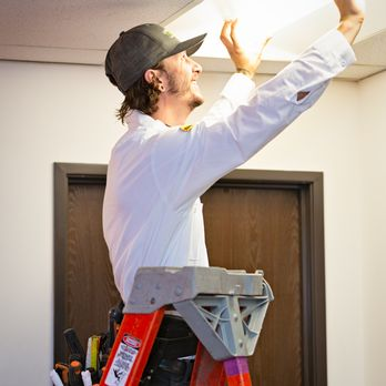 An electrician from Genesis Electric, wearing a white shirt and cap, stands on a ladder fixing a ceiling light in an indoor setting.