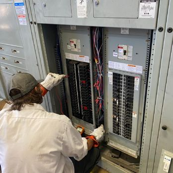An electrician working on an electrical panel, wearing a white shirt, cap, and gloves, using tools to make adjustments.