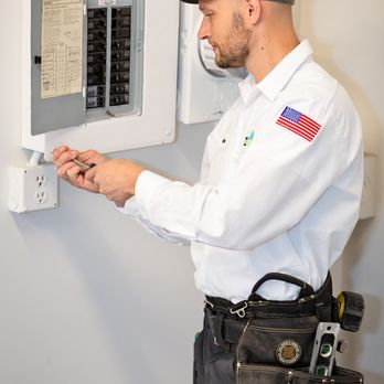 An electrician from Genesis Electric in Vallejo adjusting a circuit breaker panel, wearing a tool belt and a white uniform with a flag patch.