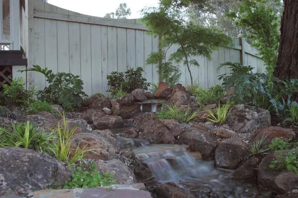 A landscaped garden in Sebastopol by Gill Landscape Inc., featuring a natural rock waterfall and lush greenery against a wooden fence.