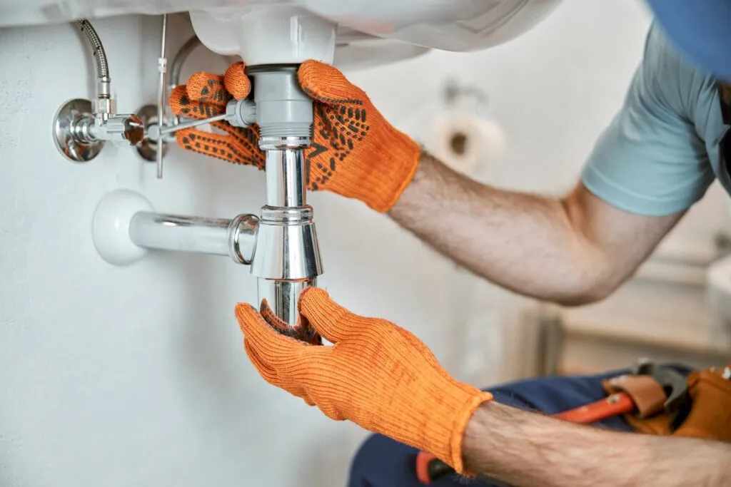 "A plumber wearing orange gloves works on a sink pipe installation, demonstrating the expertise of Gladiator Repipe, Inc. in San Jose."