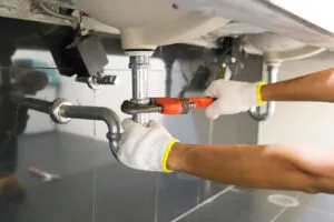 A plumber wearing gloves uses a wrench to fix pipes under a sink, representing Gladiator Repipe, Inc. in San Jose.