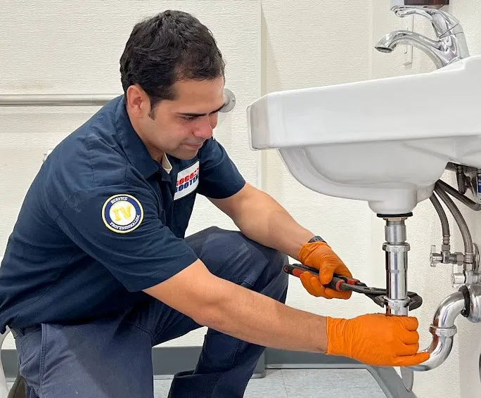 A Gogo Rooter plumber in San Jose wearing orange gloves works on a sink's plumbing with a wrench.