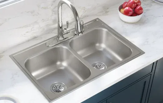A shiny stainless steel double kitchen sink with a modern faucet, set on a marble countertop, accompanied by a bowl of red apples.