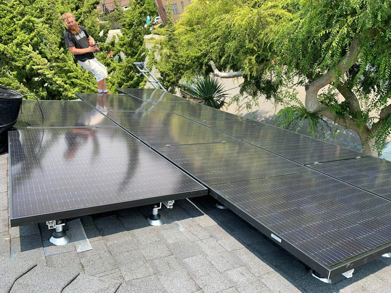 A person stands on a roof next to installed solar panels surrounded by trees.