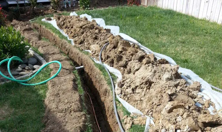Image of a freshly dug trench in a green yard, showcasing the excavation work by Gray Plumbing Inc in Fairfield, with pipes laid alongside the trench.
