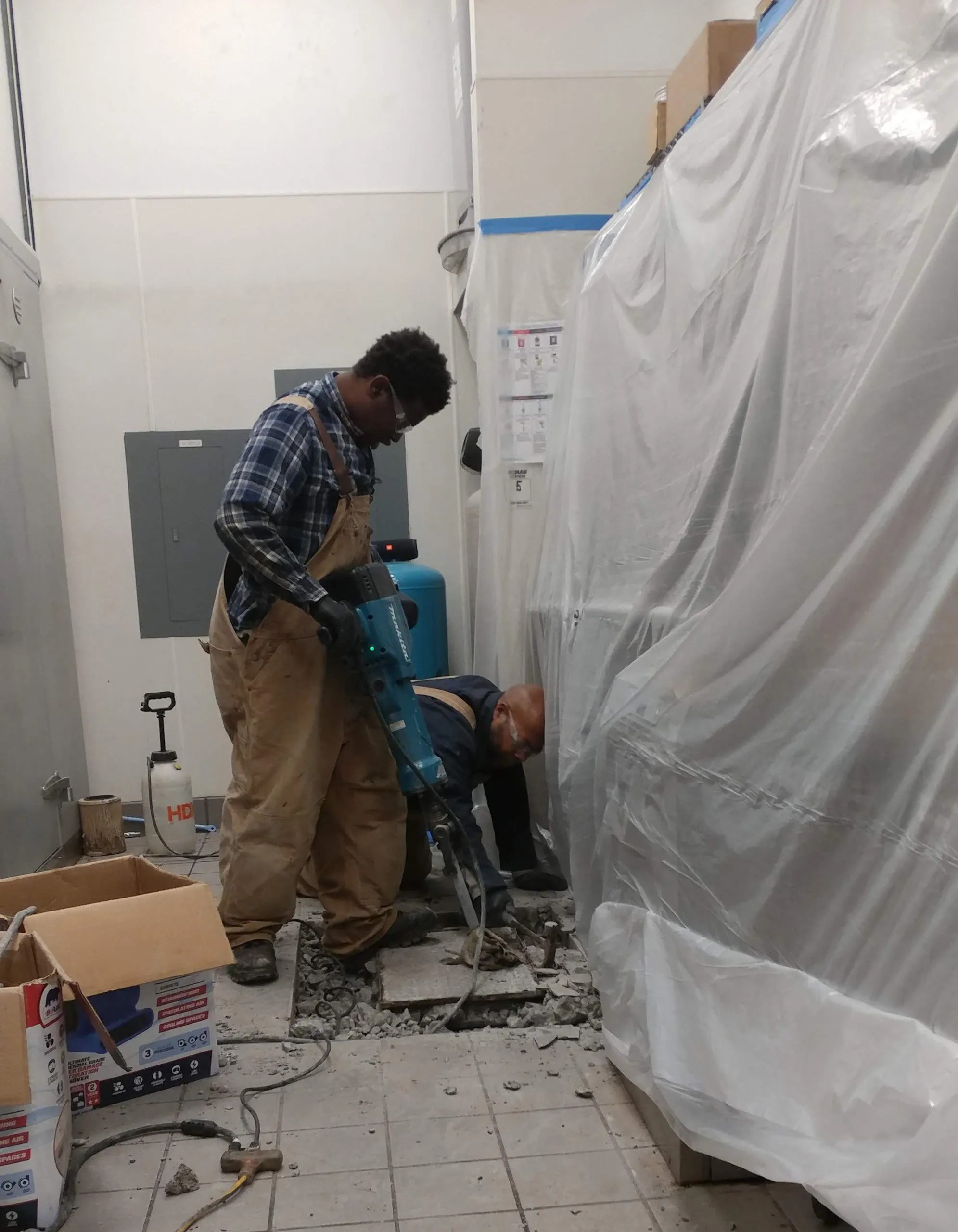 Two workers from Gray Plumbing Inc in Fairfield are using a jackhammer to break up a tiled floor, surrounded by boxes and covered equipment.