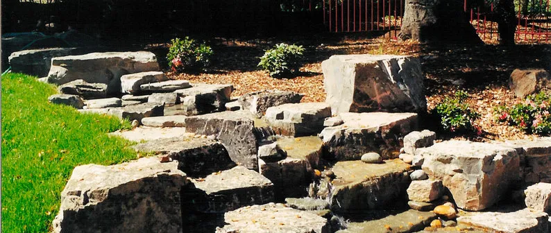 A natural stone landscape feature with a small water stream, surrounded by greenery, designed by Grimes Natural Landscape in Los Altos.