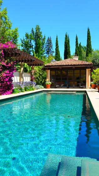 A luxurious outdoor swimming pool surrounded by lush greenery and a wooden pergola at a retreat in Los Altos.