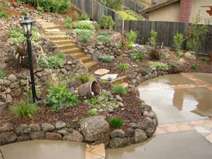 A landscaped garden with stone steps, a variety of plants, a lamppost, and a patio area, designed by Hansen Landscape Contractor in Castro Valley.