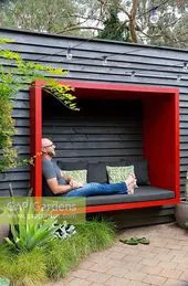 A person relaxing on a modern red-framed outdoor bench with cushions, surrounded by greenery, in a garden setting.