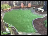 A beautifully landscaped backyard featuring lush artificial grass, surrounded by flower beds, two chairs, and a bench.
