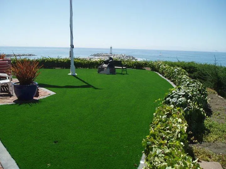 A vibrant green artificial turf lawn overlooks the ocean, bordered by hedges and potted plants, with a lighthouse visible in the background.