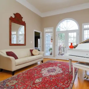 A sophisticated living room featuring a white piano, a cream-colored sofa with decorative pillows, a large mirror above the sofa, and a vibrant red patterned rug with natural light streaming through arched windows.